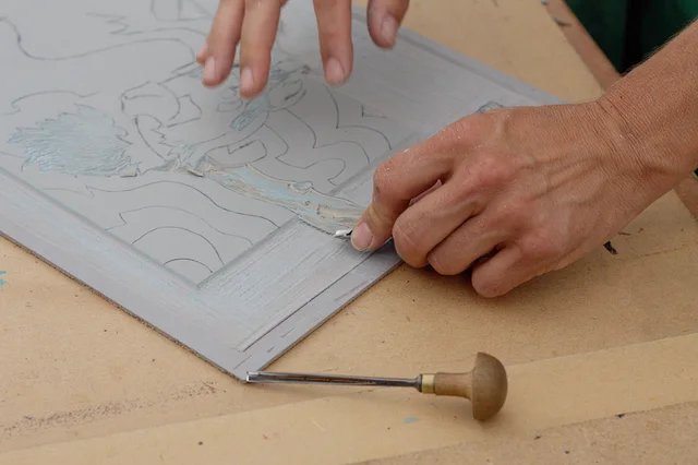 Hands using a gouging tool to carve a design into a linoleum block.