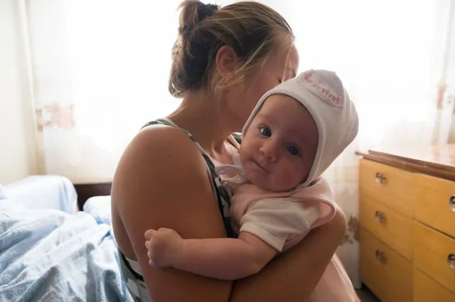 A mother holding her baby beside a sunlit window.